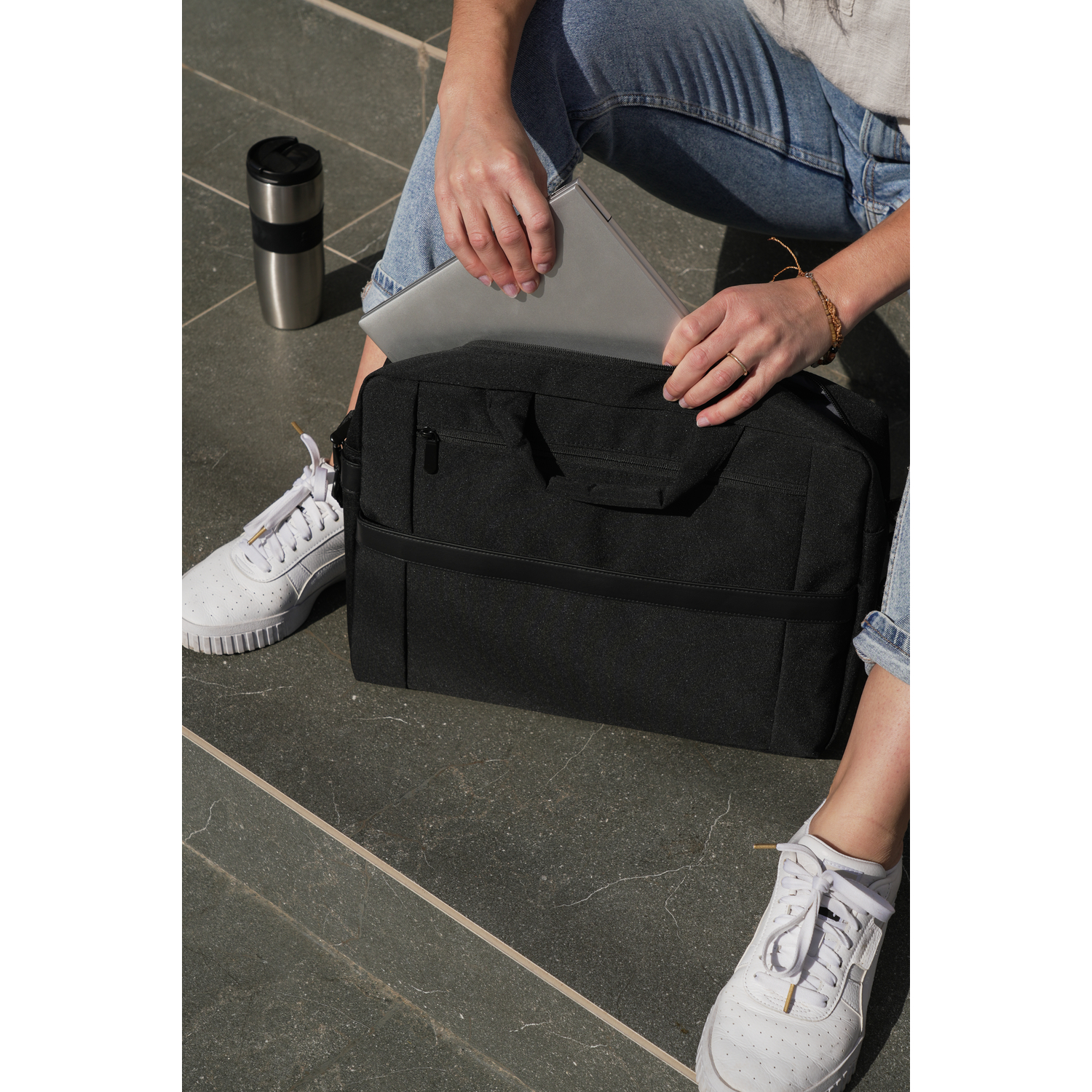 Woman putting a laptop into a black bag. There's a silver thermos and white sneakers on the ground.