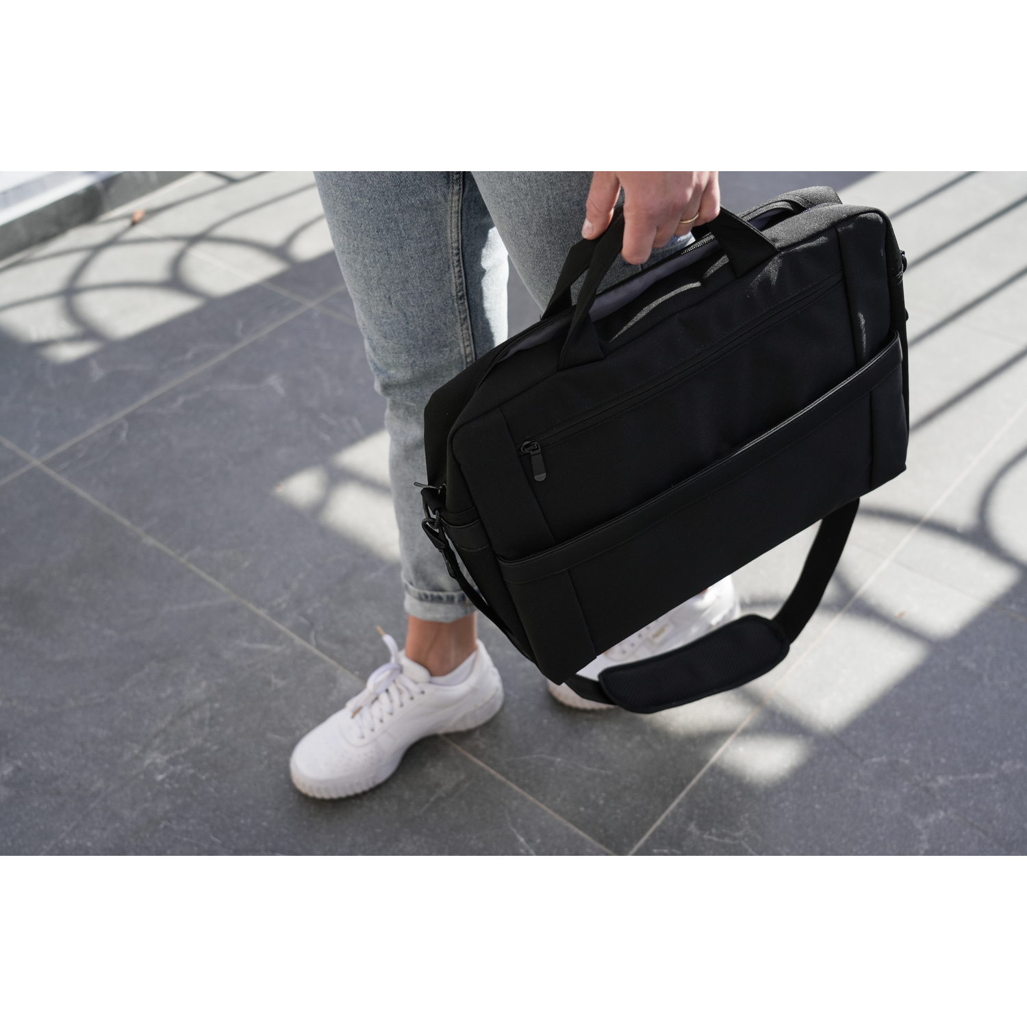 Person holding a black briefcase with a shoulder strap, walking on a gray surface.