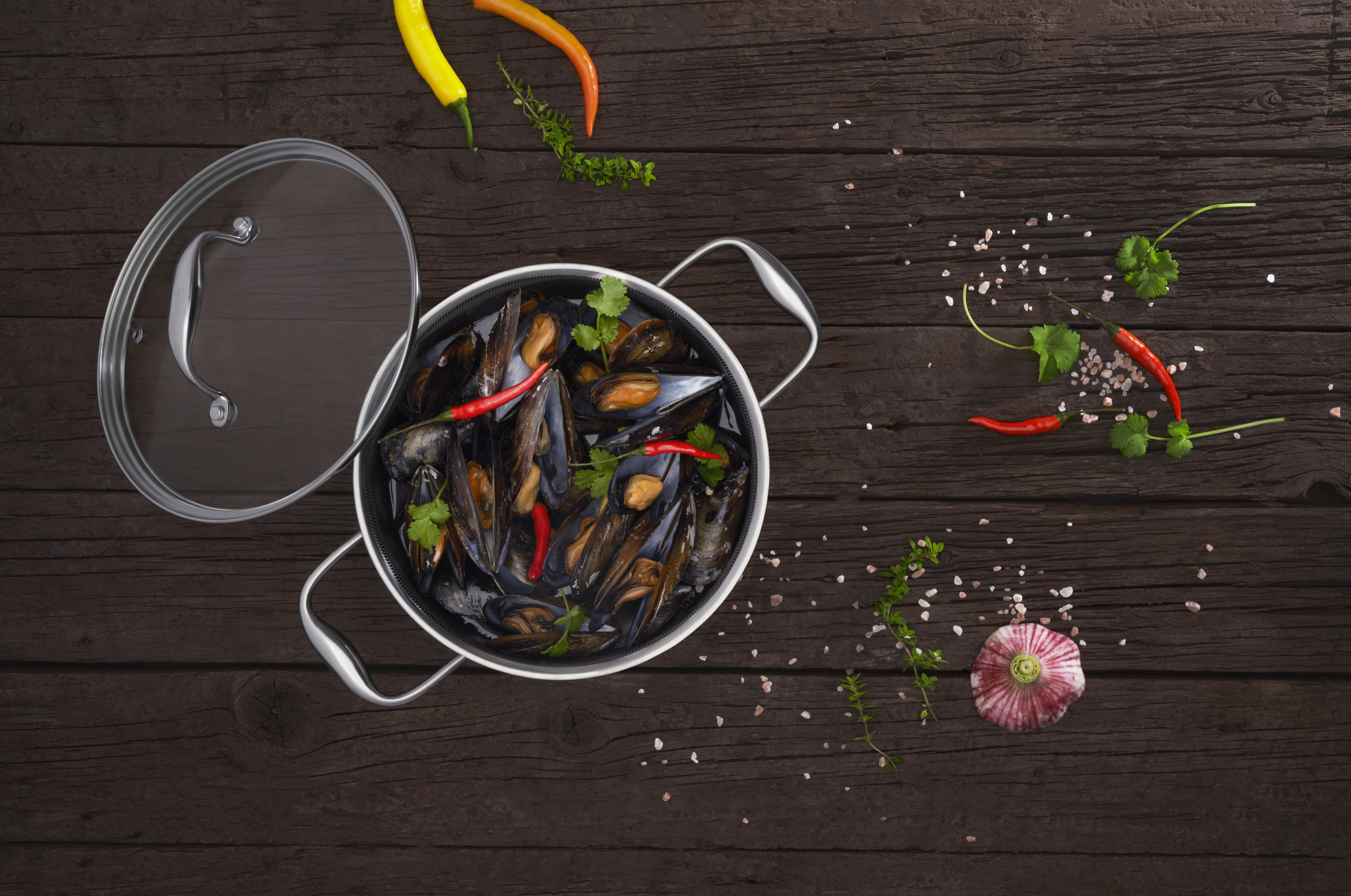 Widok z góry na małże w garnku, z papryczkami chili i ziołami. Ciemny drewniany stół.
