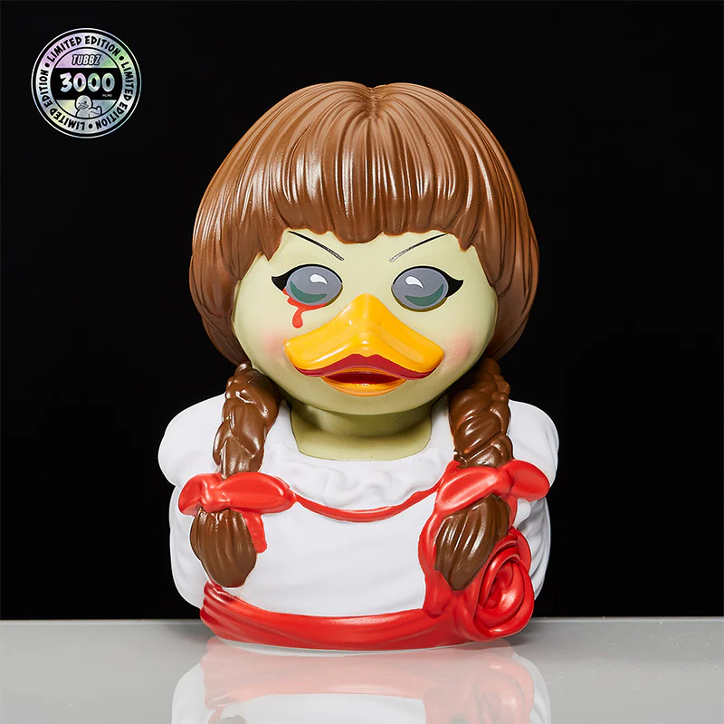 Annabelle kacsa alakú figura, barna hajjal, fehér ruhával, piros szalagokkal.