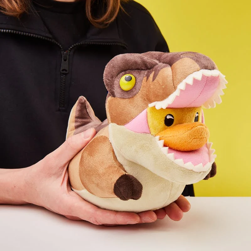 Dinoszaurusz-kacsa plüssjáték kézben, sárga háttéren.