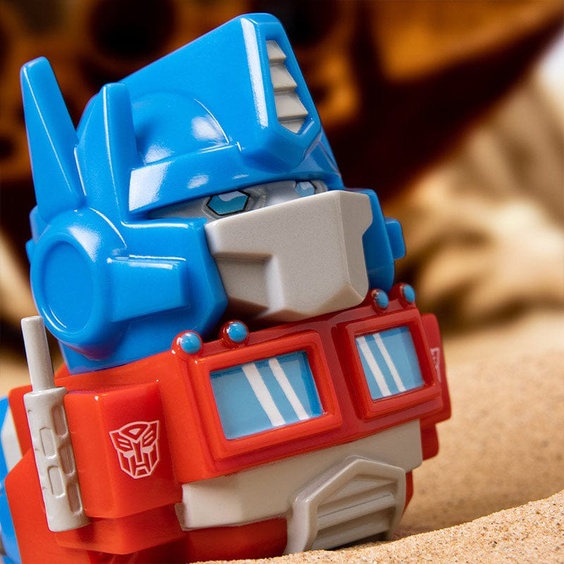 Közeli kép egy kék és piros Optimus Prime játékfejről homokon. Egy piros Autobot szimbólum látható.