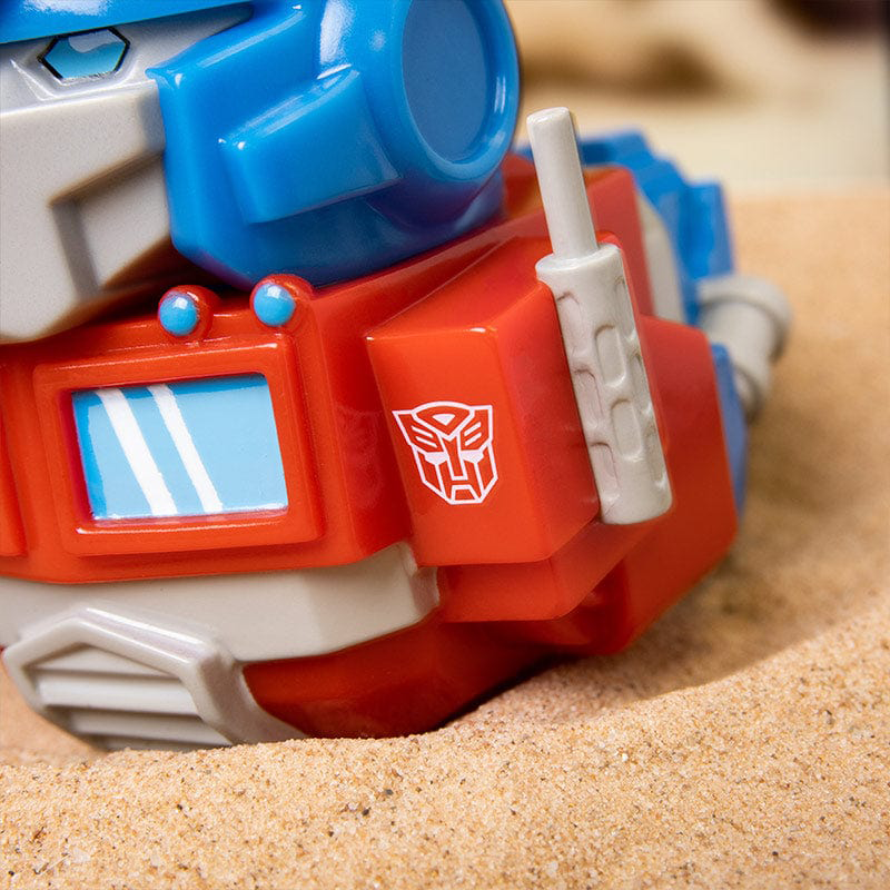 Egy piros-kék robot Transformers játék homokon, fehér emblémával.