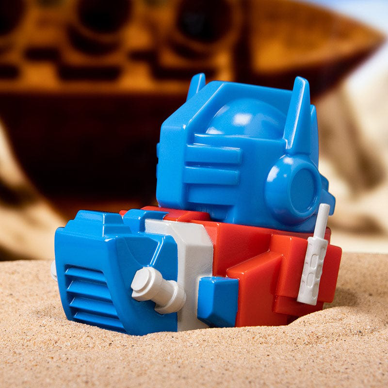 Kék és piros Transformer játék a homokban, homályos hajó háttérrel.