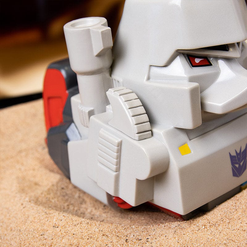 Megatron játékfejének közeli képe homokon, a test egy részével.