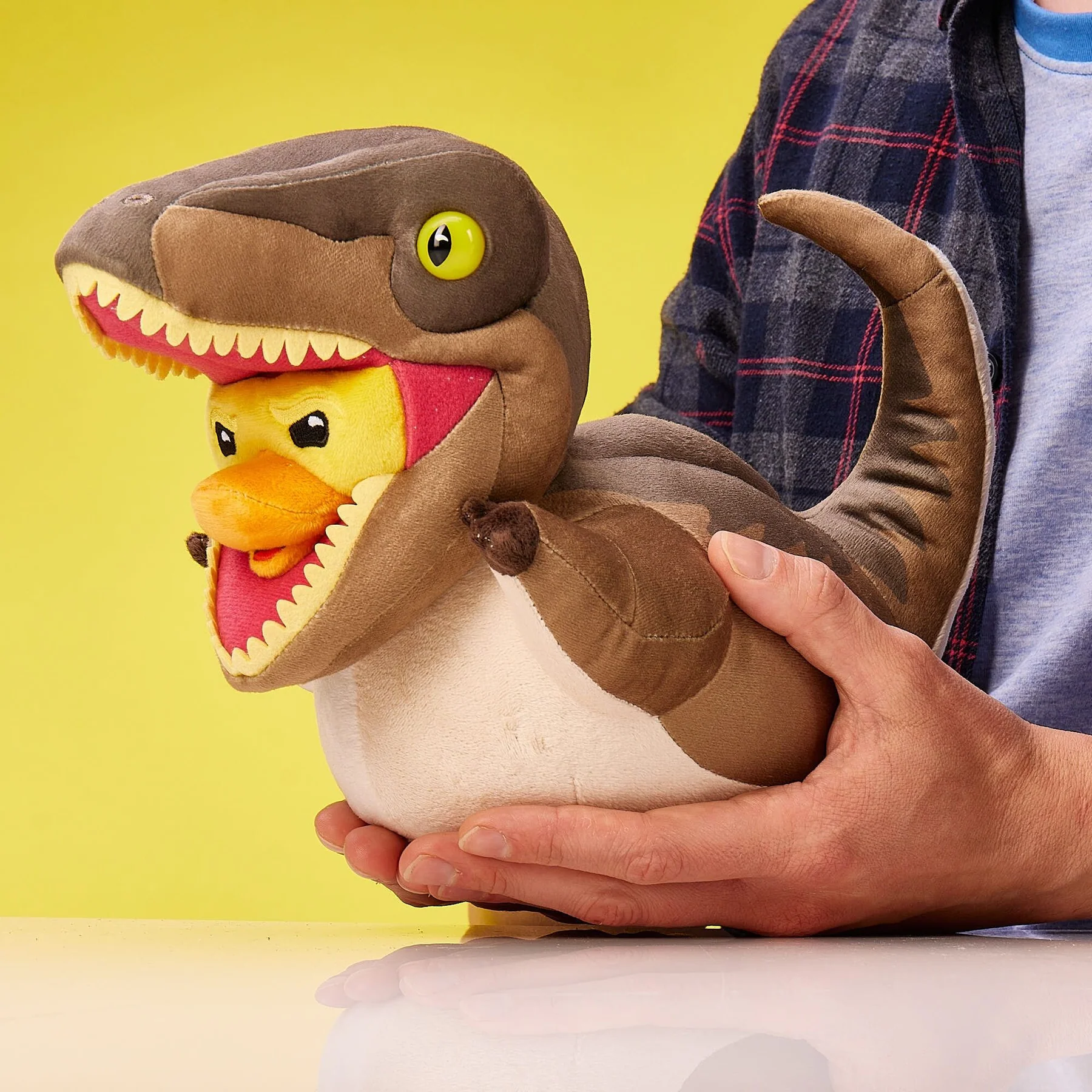 Egy személy egy dinoszaurusz plüssjátékot tart, szájában egy kacsával, sárga háttér előtt.
