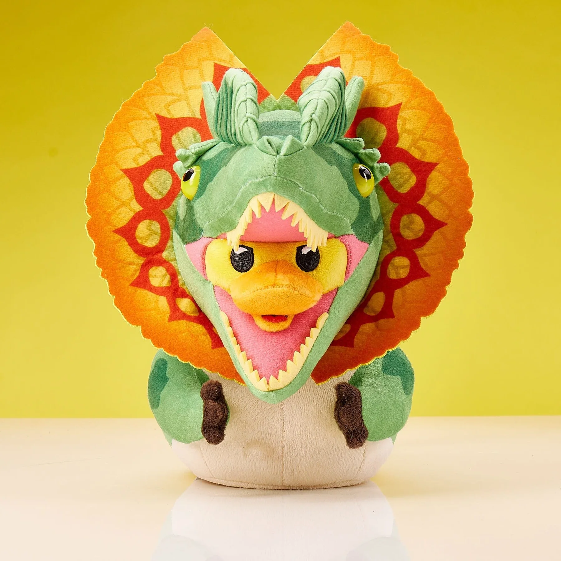 Zöld és narancssárga Dilophosaurus plüssjáték nyitott szájjal. Sárga háttér előtt.