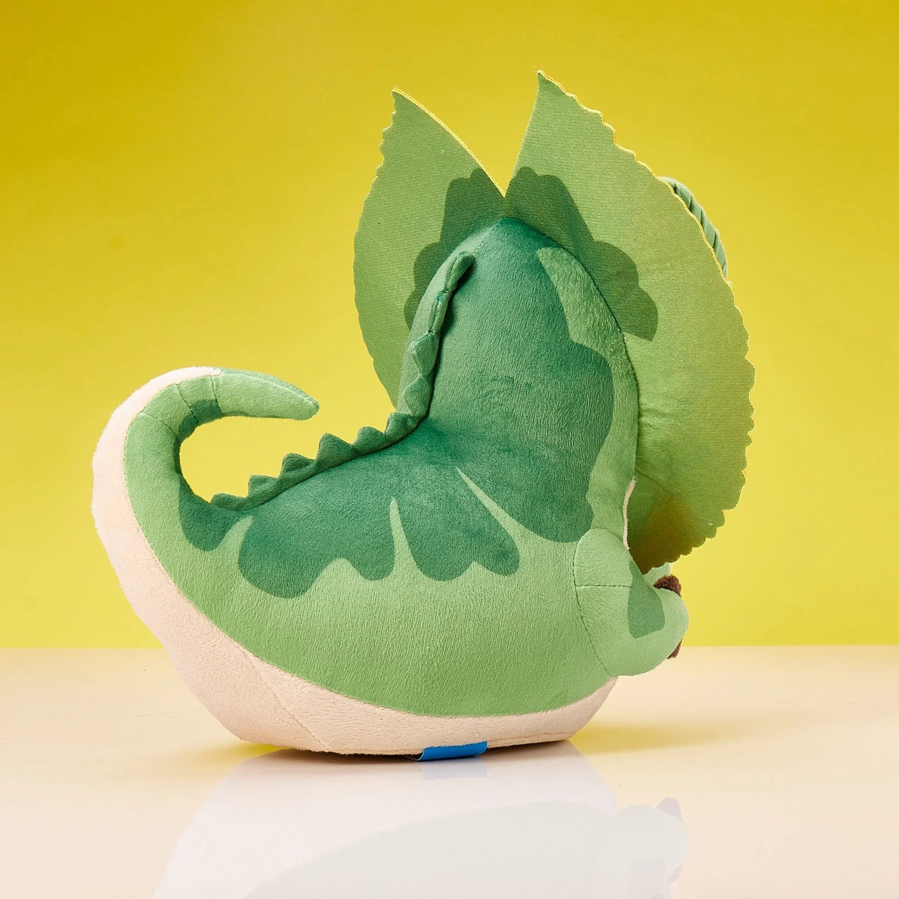 Zöld plüss dinoszaurusz nagy fodrokkal, oldalnézet. Sárga háttér, fehér felület.