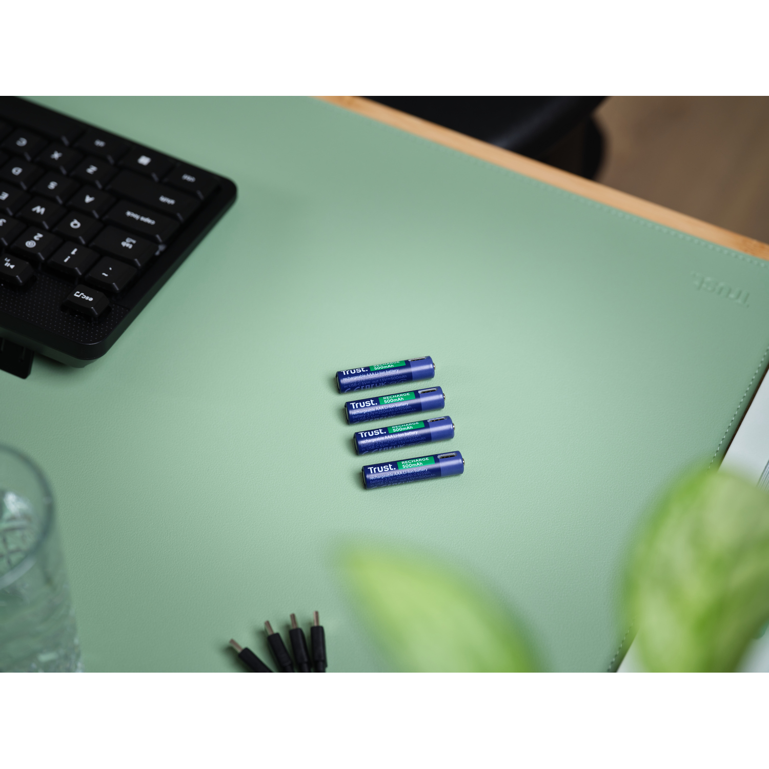 Vier Trust AA-batterijen liggen op een lichtgroen bureau. Een toetsenbord, een glas en een plant staan op het bureau.