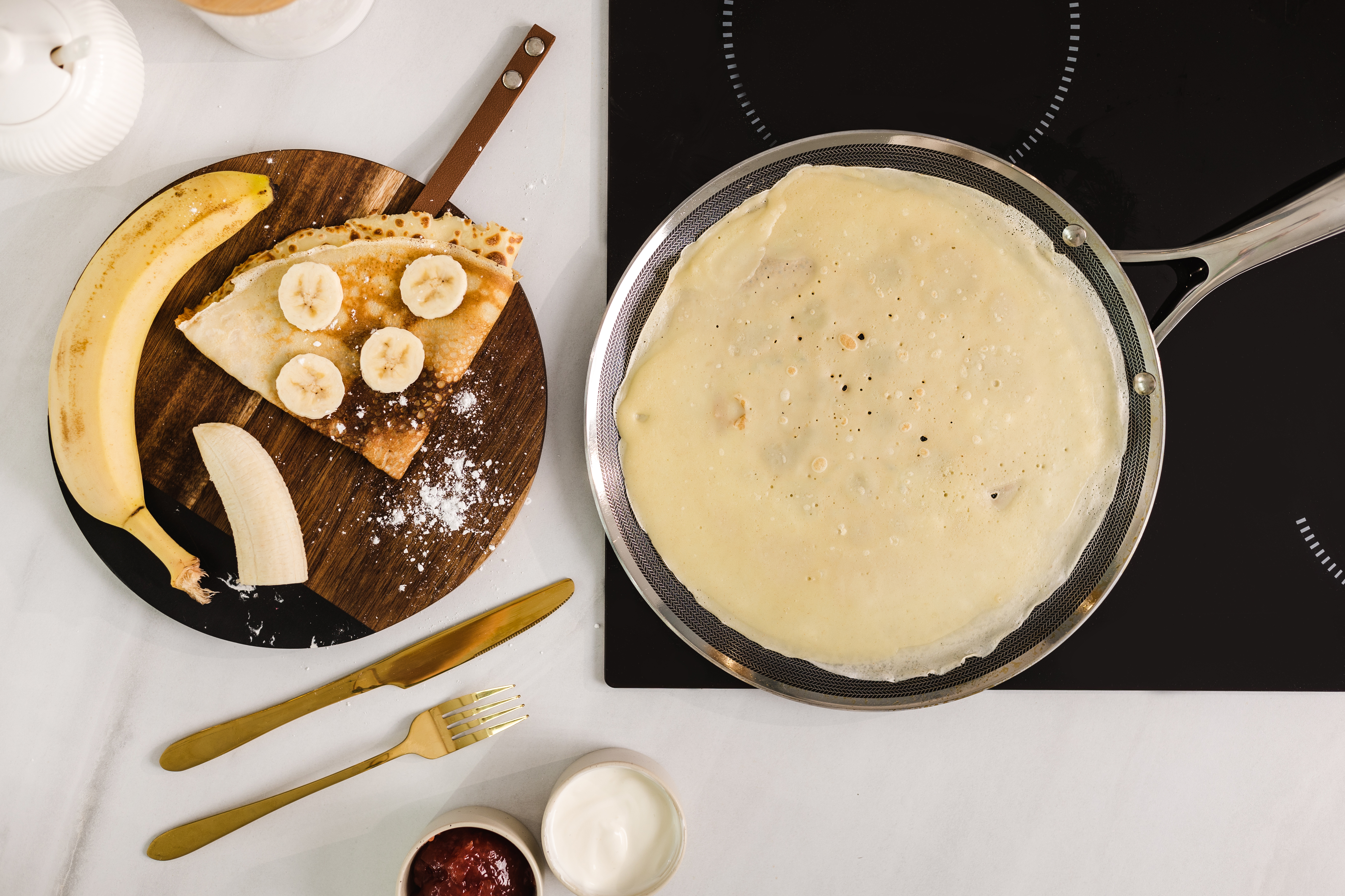 Naleśnik na patelni, z bananami i cukrem pudrem na drewnianej desce, obok sztućców.