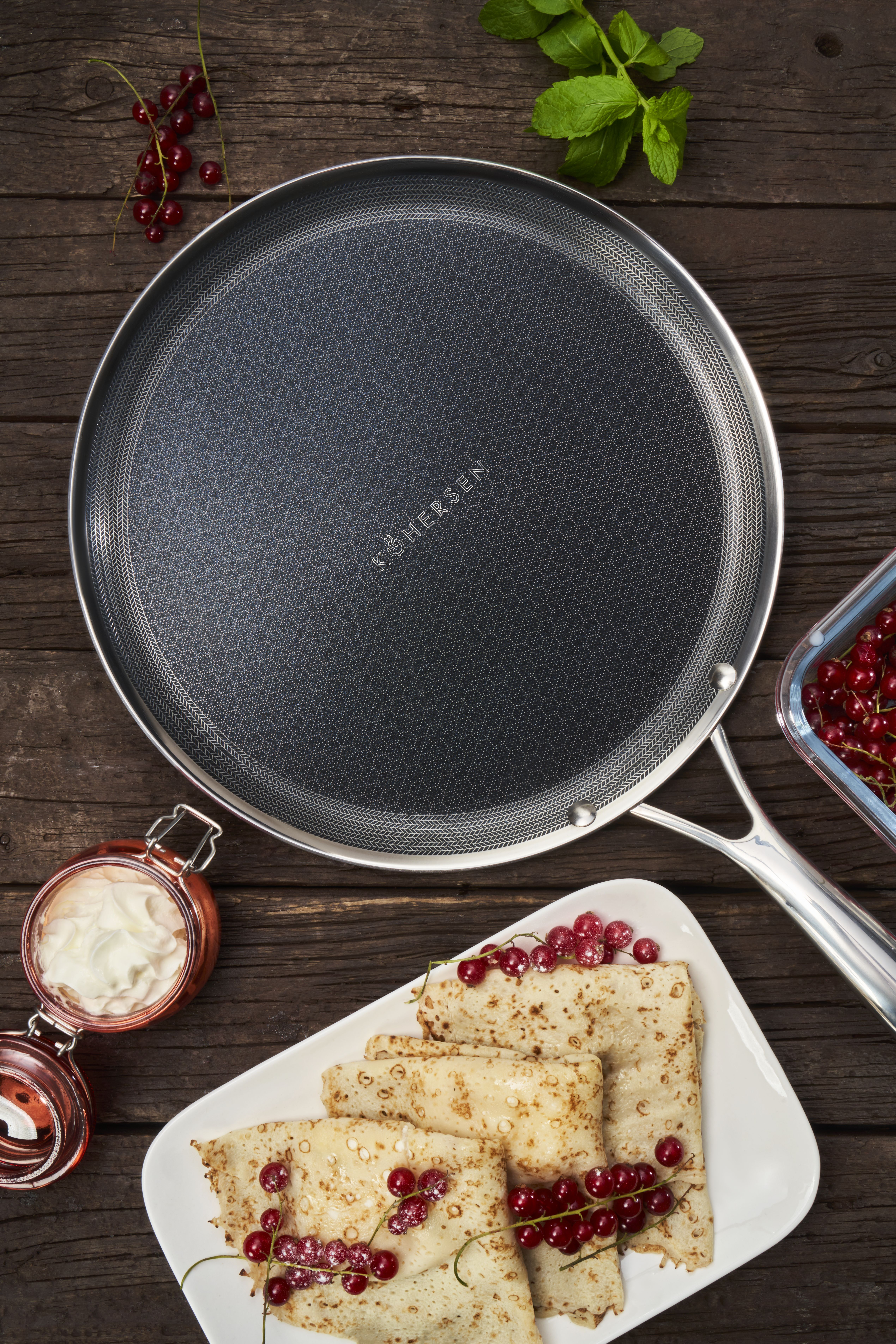 Patelnia z naleśnikami, czerwonymi jagodami i śmietaną na drewnianym stole. Liście mięty z boku.