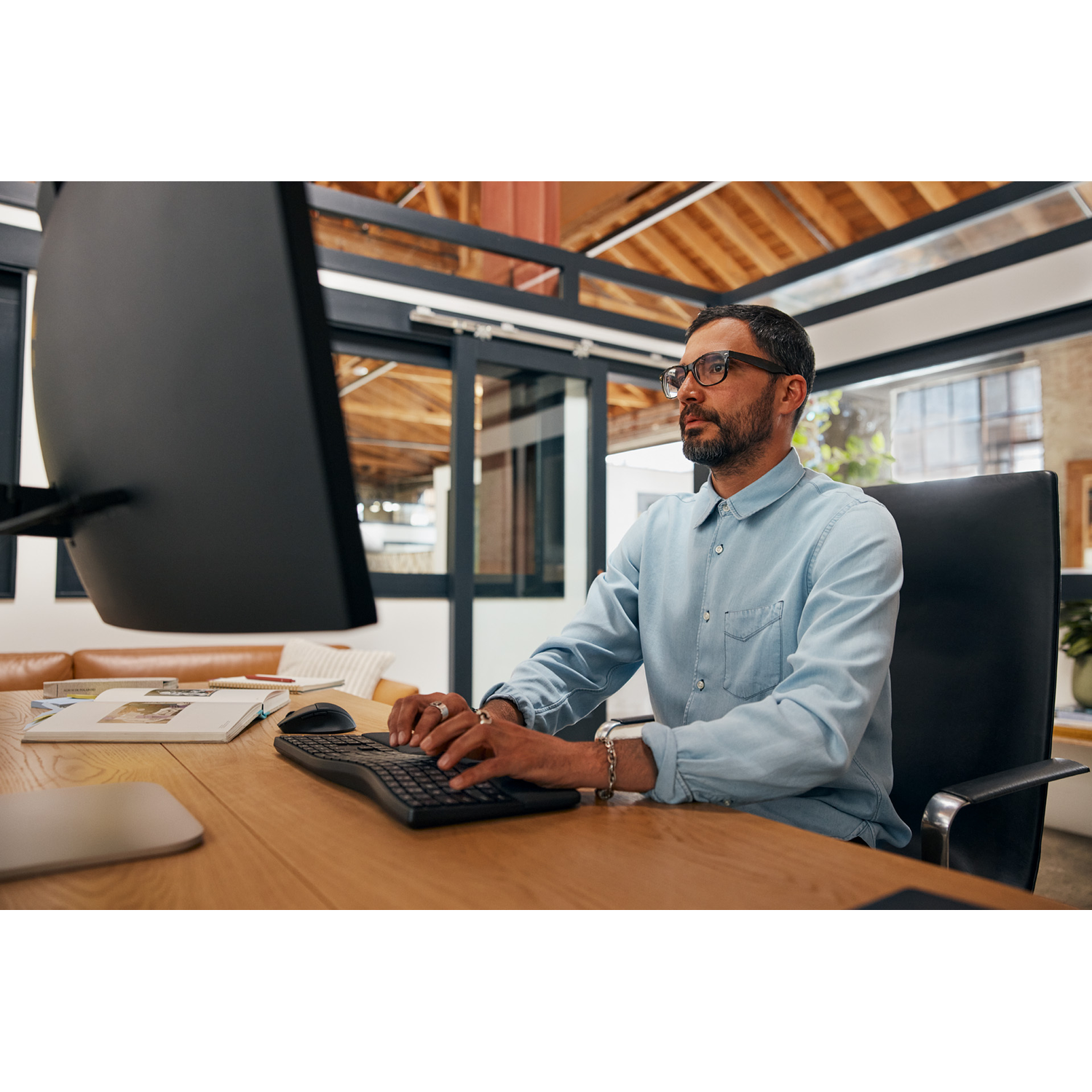 Man with glasses working on a computer, wooden desk, office setting.
