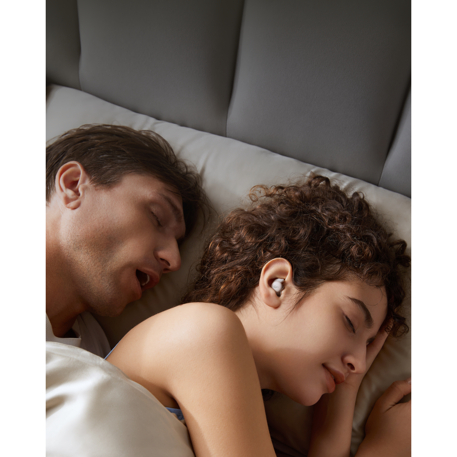 A couple sleeping in bed. The woman has ear buds in. The background is dark.