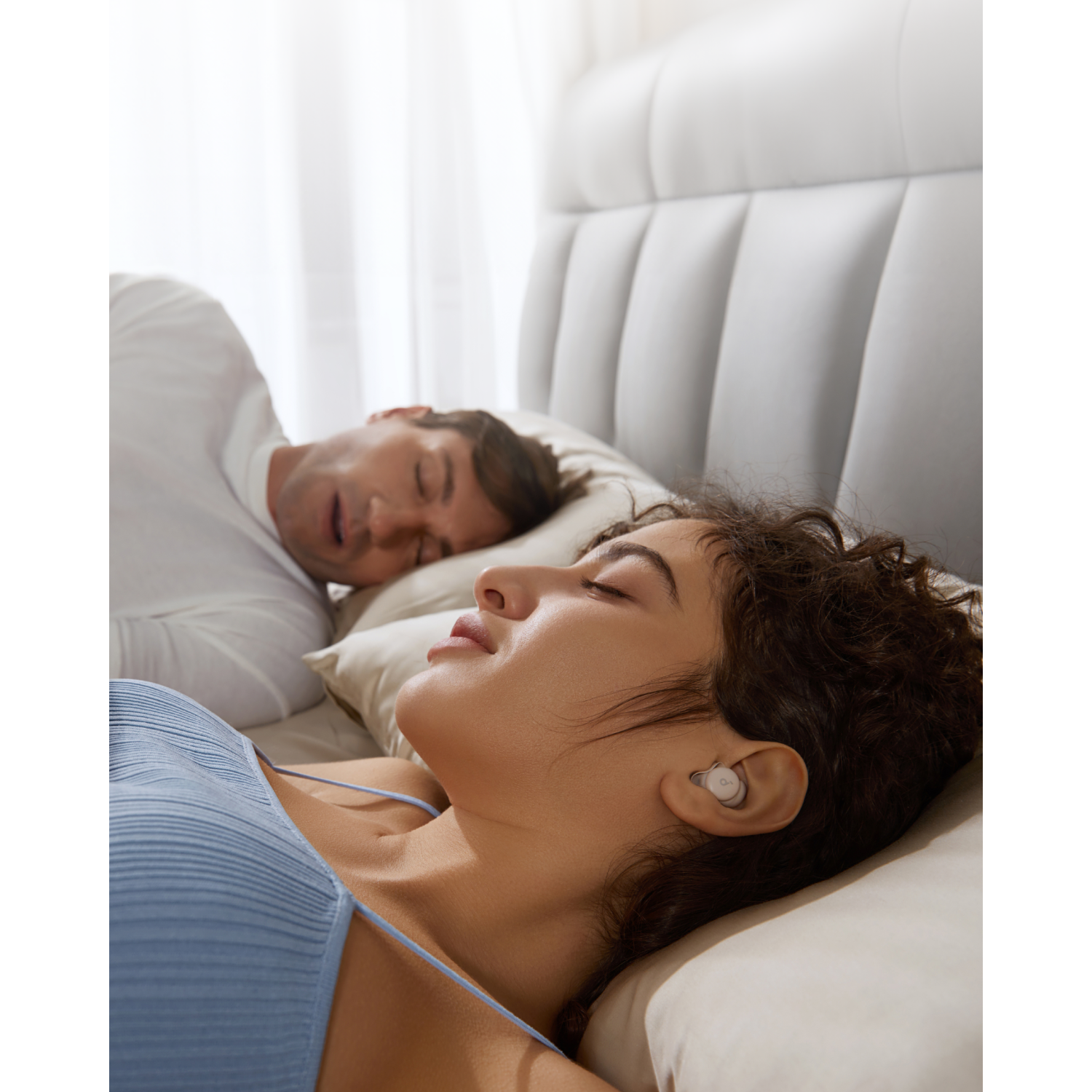 A woman sleeps with an earbud in, next to a man sleeping in a bed.