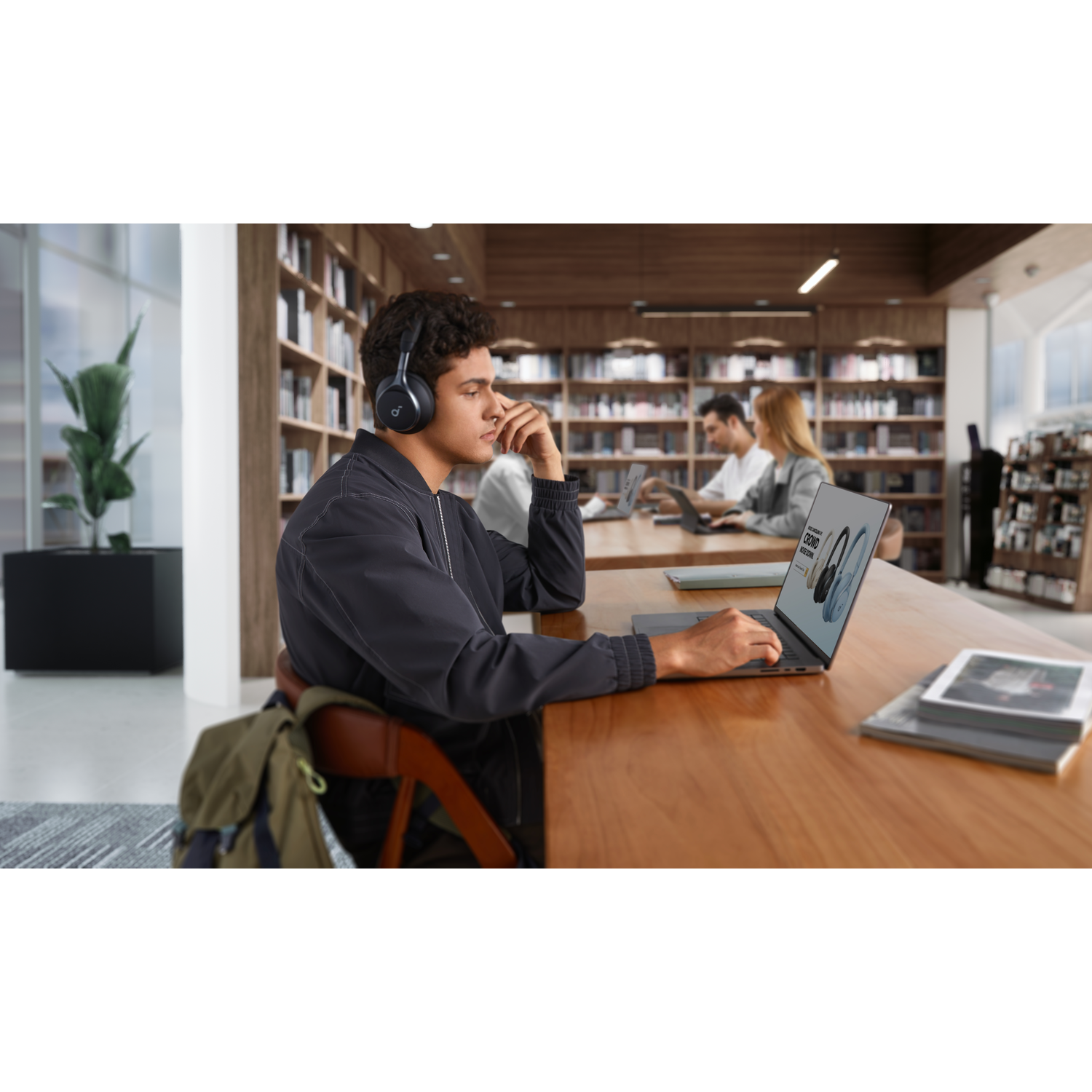 A person wearing headphones, using a laptop in a library setting. Other people are visible.