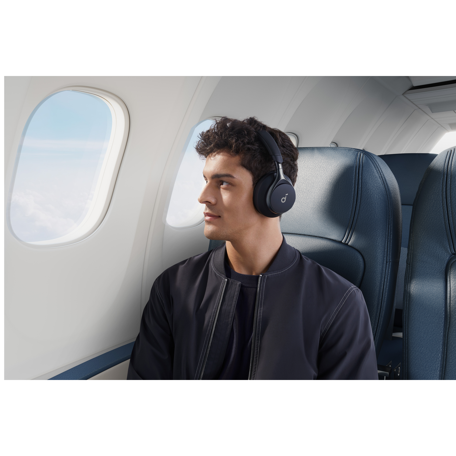 A man wearing noise-canceling headphones looks out a plane window, with clouds visible.