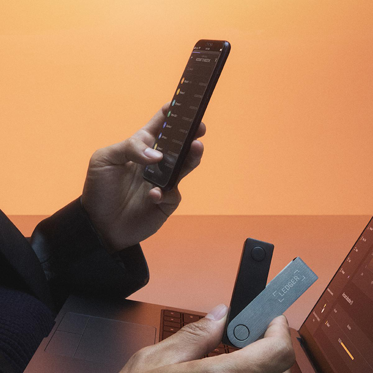 Person hält ein Telefon und Ledger Hardware Wallets, im Hintergrund ein Laptop.