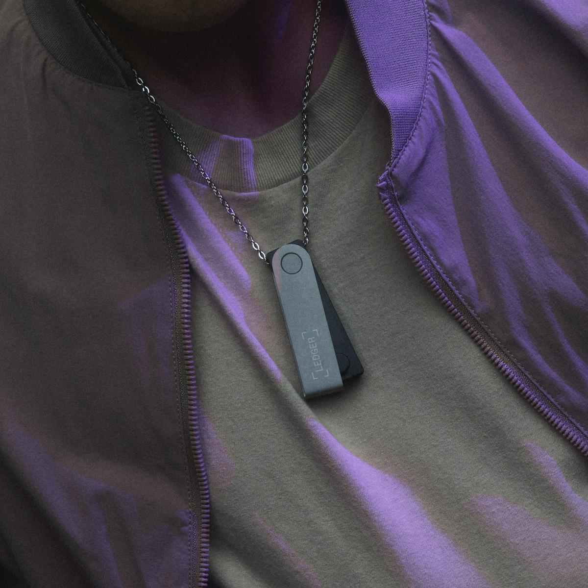 Eine Person trägt eine silberne und schwarze Ledger Krypto-Hardware-Wallet-Halskette. Dezente lila Beleuchtung.