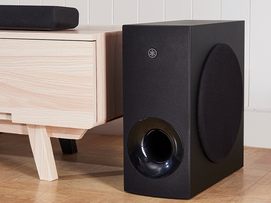 Czarny subwoofer i drewniana szafka z listwą głośnikową na górze. Podłoga jest drewniana.