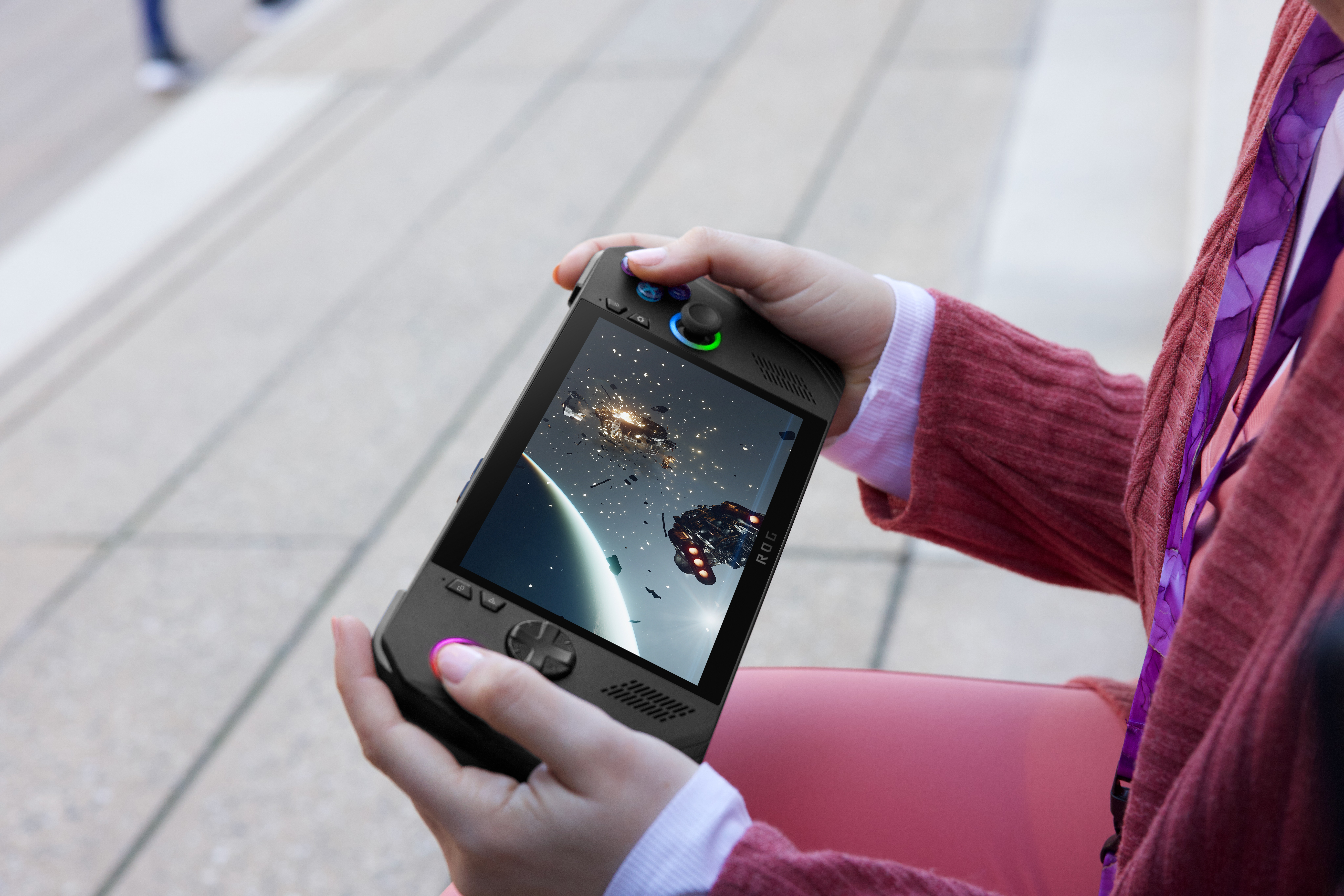 Eine Person hält ein tragbares Spielgerät. Der Bildschirm zeigt eine Weltraumschlachtszene mit Schiffen und Explosionen.