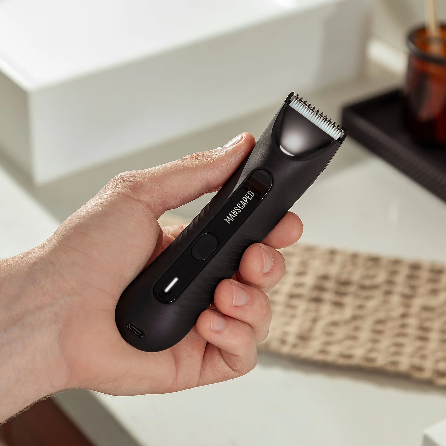 Eine Hand, die einen schwarzen Manscaped-Trimmer hält. Ein weißes Waschbecken und eine beige Matte sind im Hintergrund.