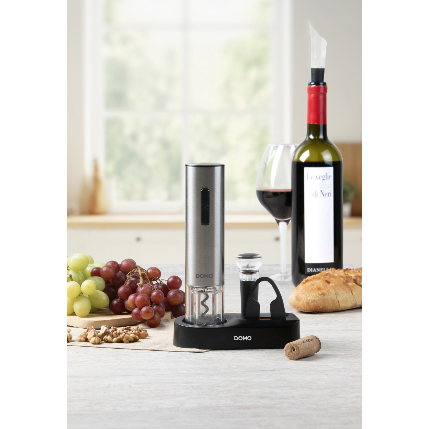 A wine bottle, a corkscrew, a glass of red wine, grapes, and a loaf of bread on a table.