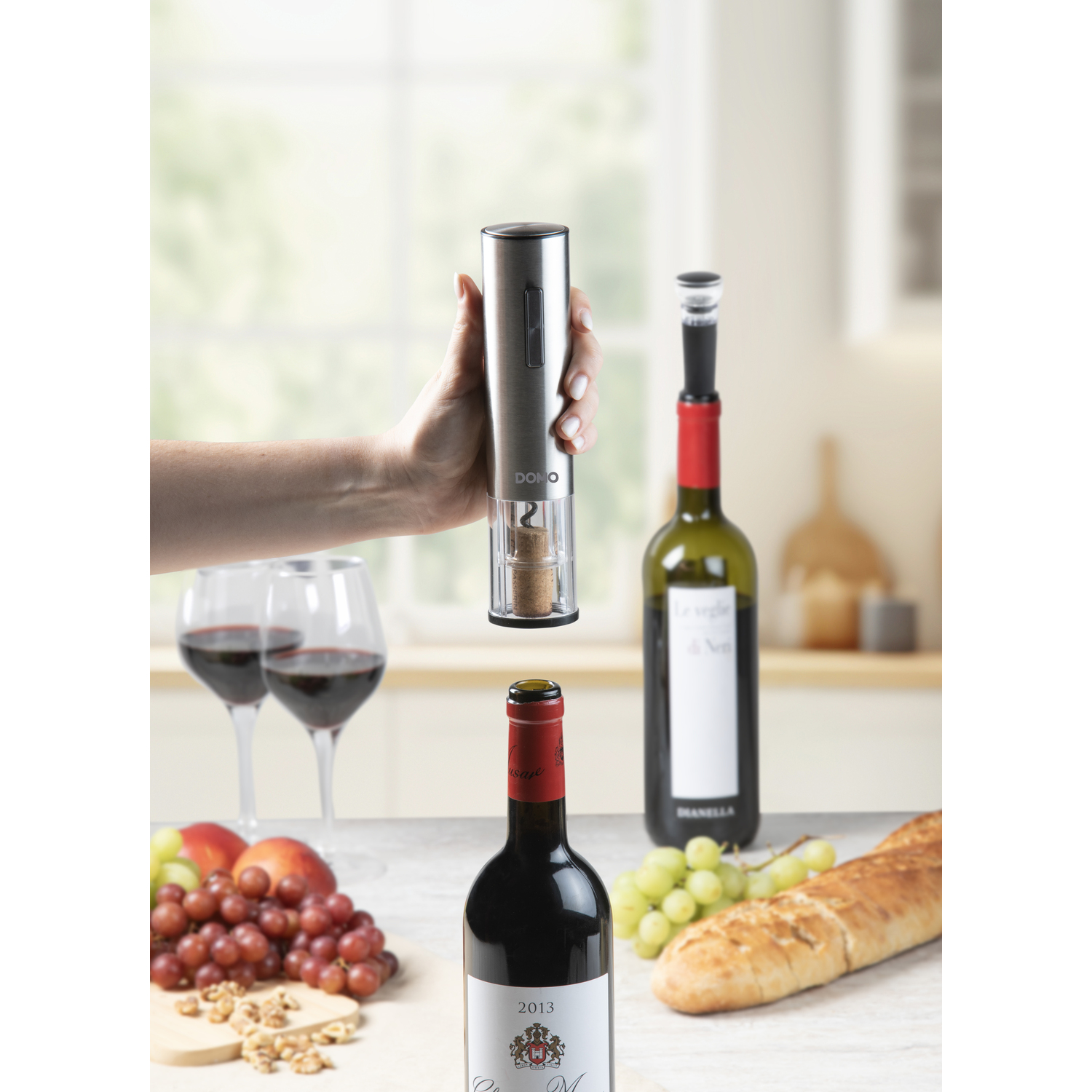 A person opening a wine bottle with an electric opener. Two wine glasses and a red wine bottle.
