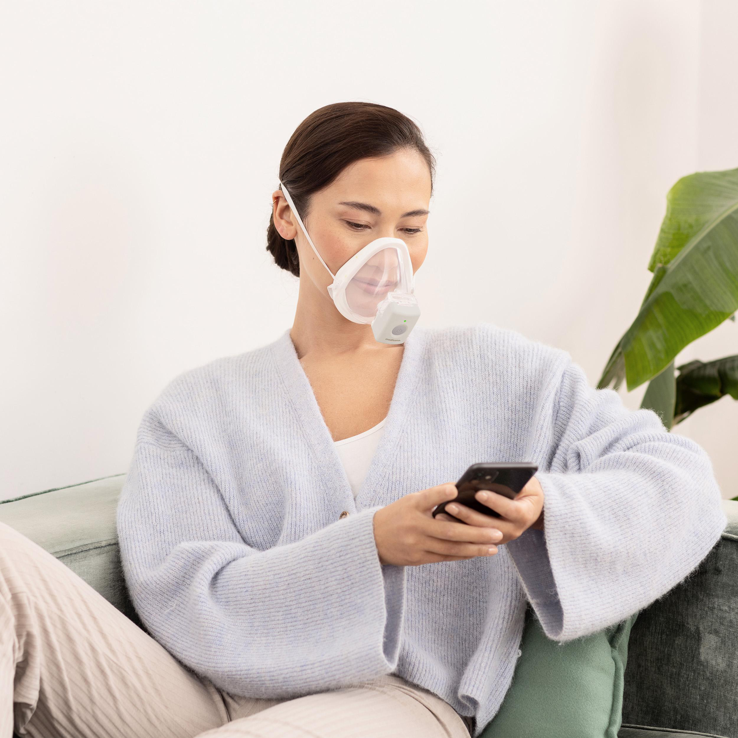 Frau mit Maske, Handy nutzend, auf Sofa sitzend.