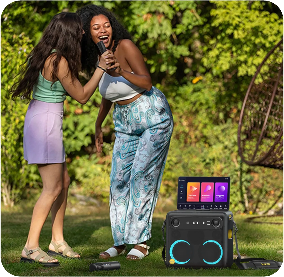Zwei Frauen singen im Freien Karaoke. Eine hält ein Mikrofon, die andere lächelt. Vorne steht eine Karaoke-Maschine.