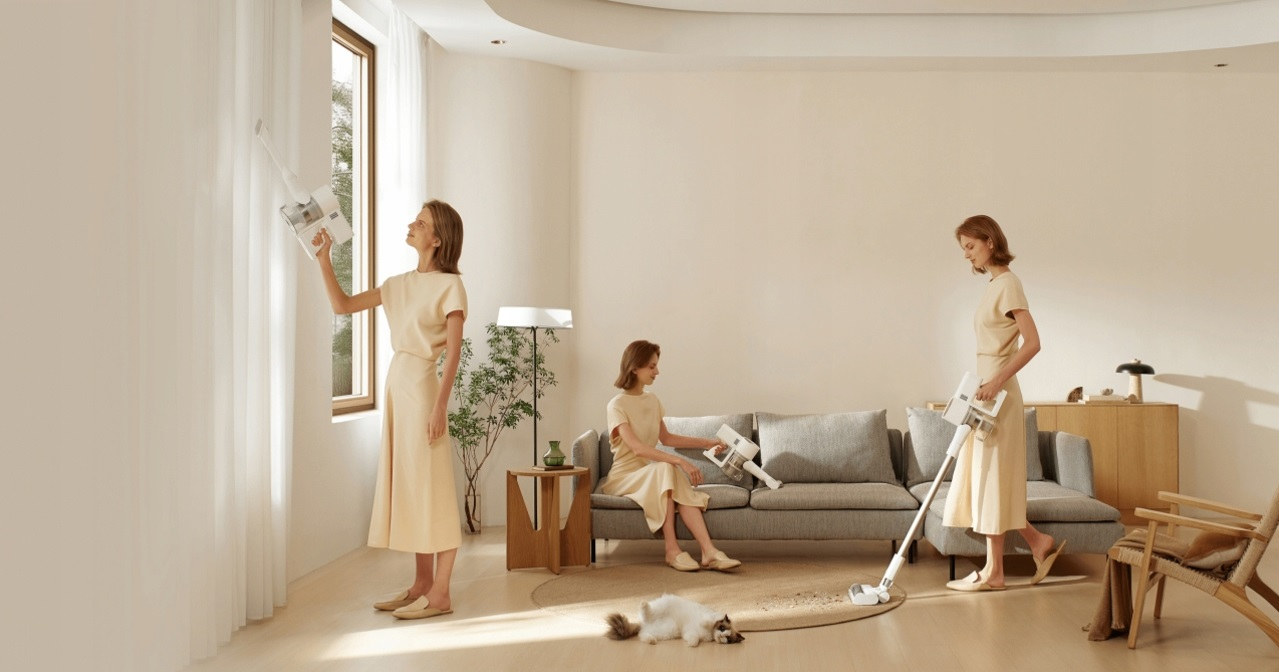 Kobieta odkurza przy oknie, na sofie i na podłodze. Kot odpoczywa na dywanie.