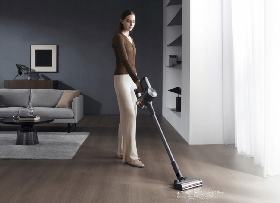 Kobieta odkurza drewnianą podłogę. Ma na sobie brązową górę i beżowe spodnie.