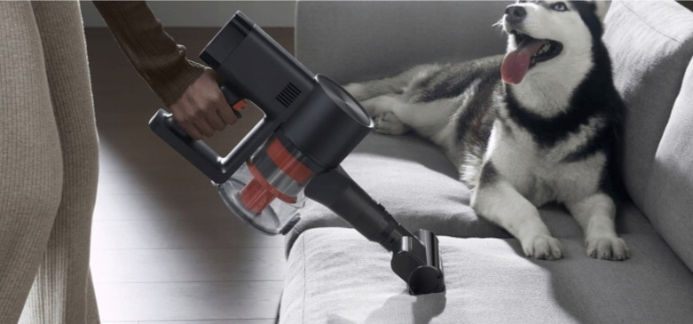 Osoba odkurza kanapę odkurzaczem bezprzewodowym. Pies leży na kanapie.