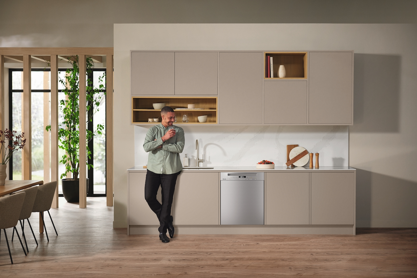 Mann in der Küche hält Glas in der Nähe eines Geschirrspülers. Beige Küchenschränke. Weiße Theke.