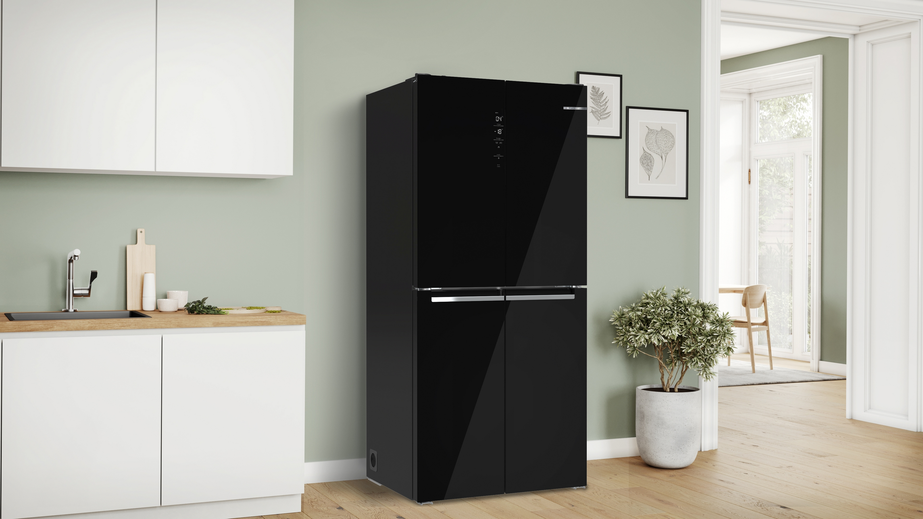 Un frigorifero nero è in una cucina con mobili bianchi e pareti verde salvia. Una pianta e arte incorniciata sono visibili.