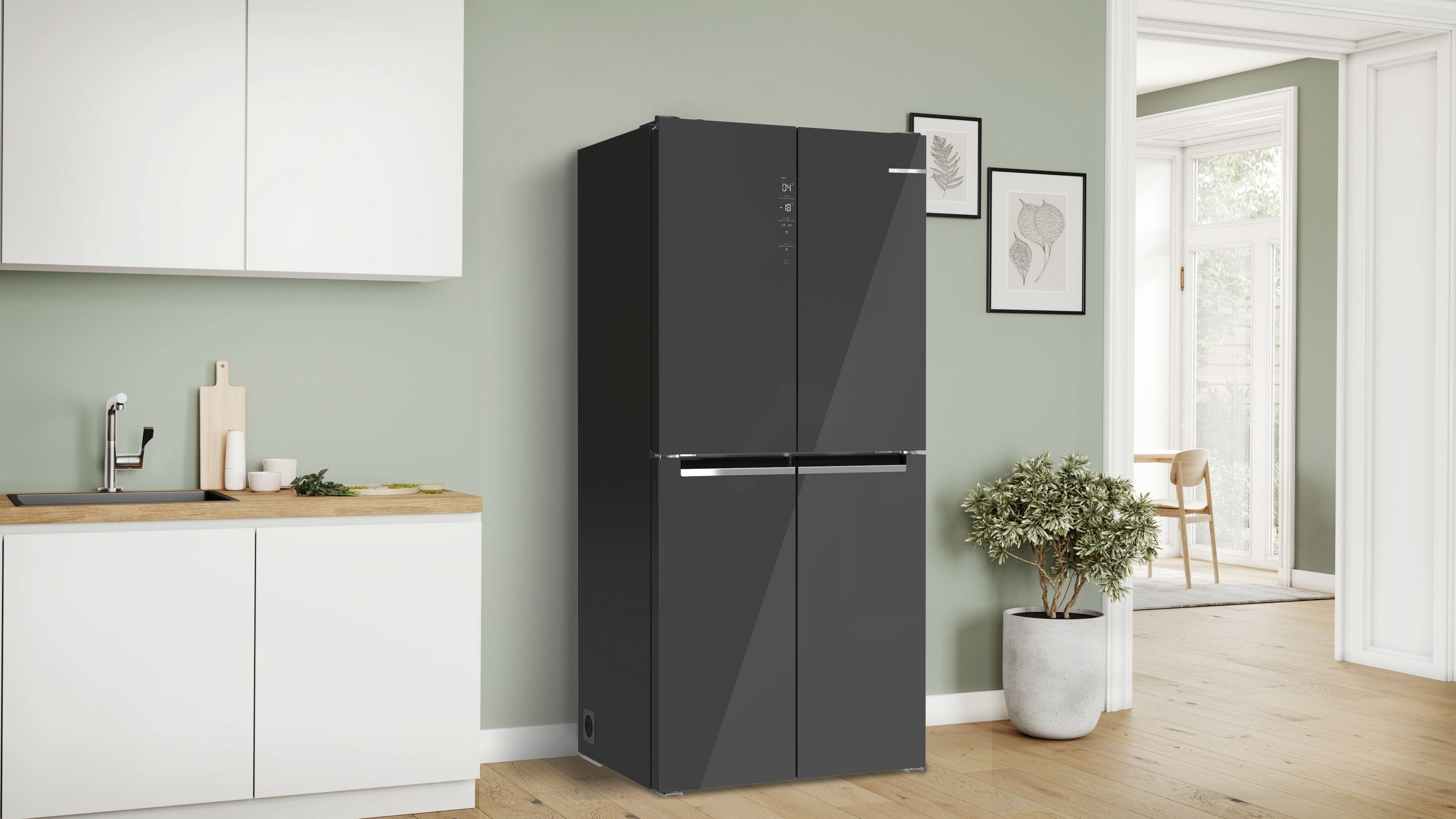 Un frigorifero nero in una cucina con mobili bianchi. Il muro è verde chiaro, c'è una pianta e arte incorniciata.