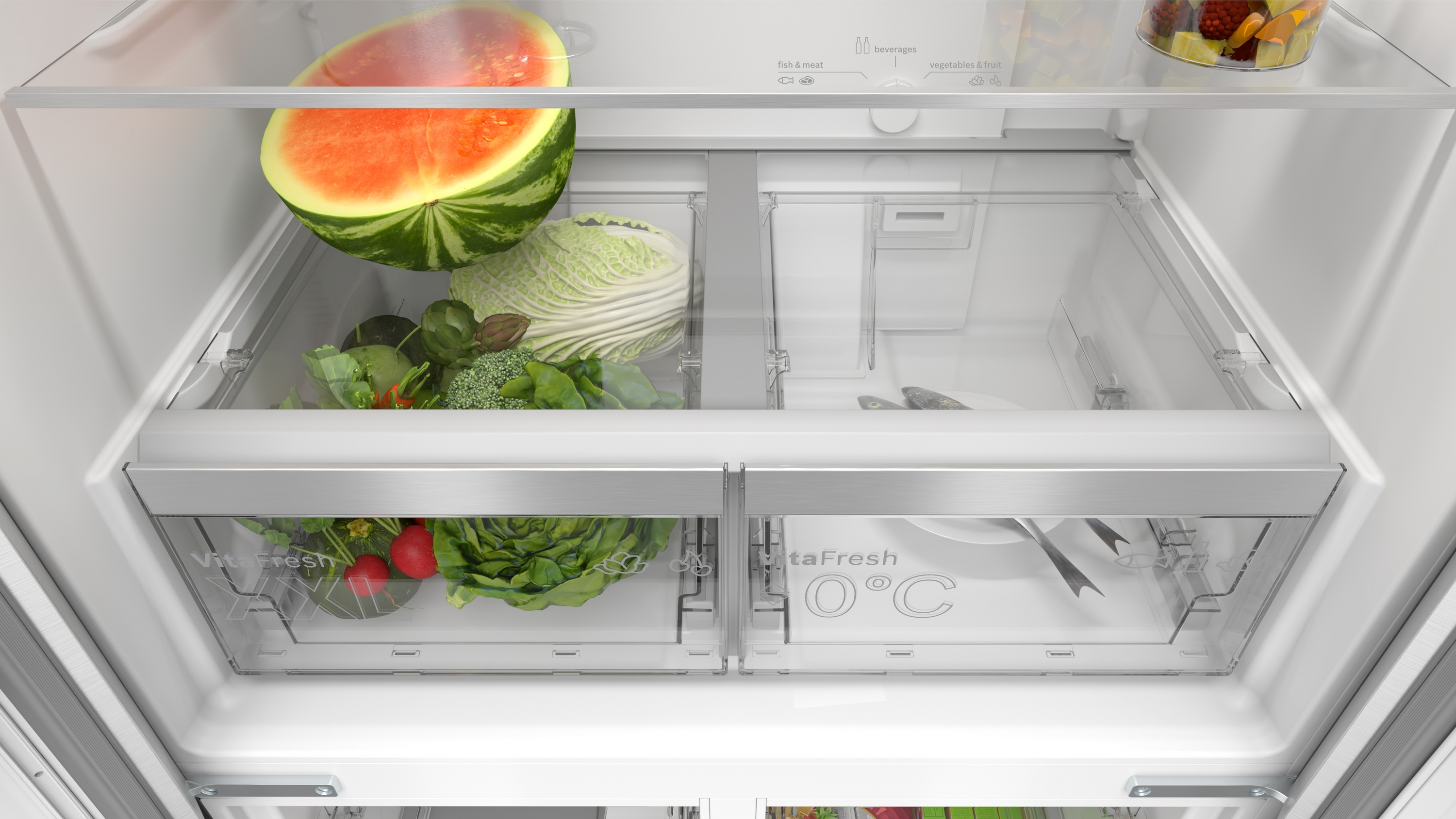 All'interno di un frigorifero: anguria, cavolo, insalata, broccoli, pesce e frutta. Cassetti trasparenti contengono cibo. Il frigo è bianco.