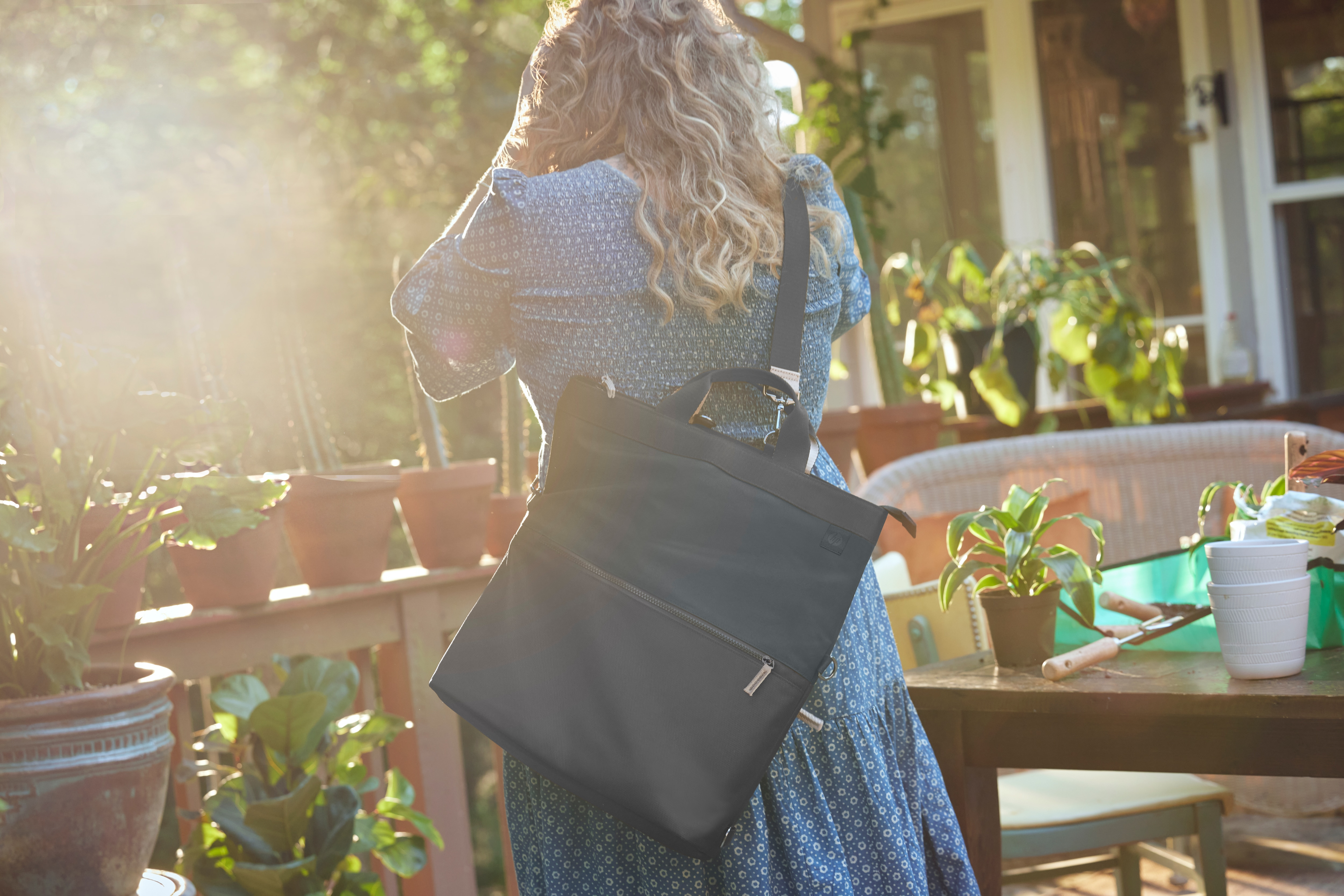 Une femme avec un sac noir sur l'épaule est dans un jardin avec des plantes en arrière-plan.