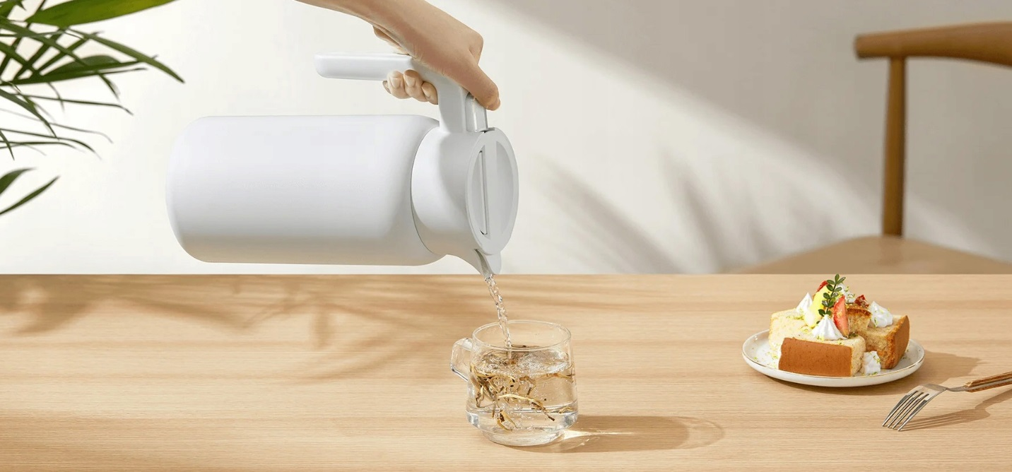 Osoba nalewa wodę z białego czajnika do szklanki. Ciasto i widelec na drewnianym stole.