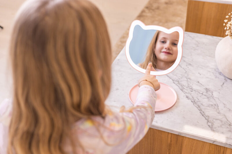 Dziewczynka patrzy w różowe lustro w kształcie kota, dotykając go palcem.