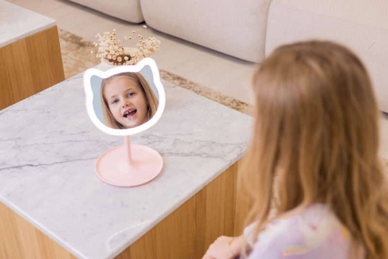 Dziewczynka patrząca na różowe lustro w kształcie kota na stole z marmurowym blatem.