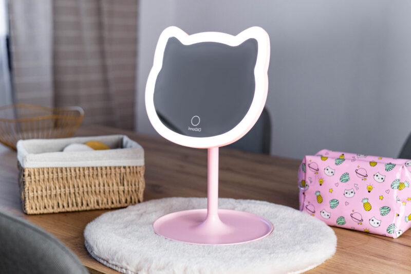 Różowe lustro w kształcie kota na stole z koszem i kosmetyczką.