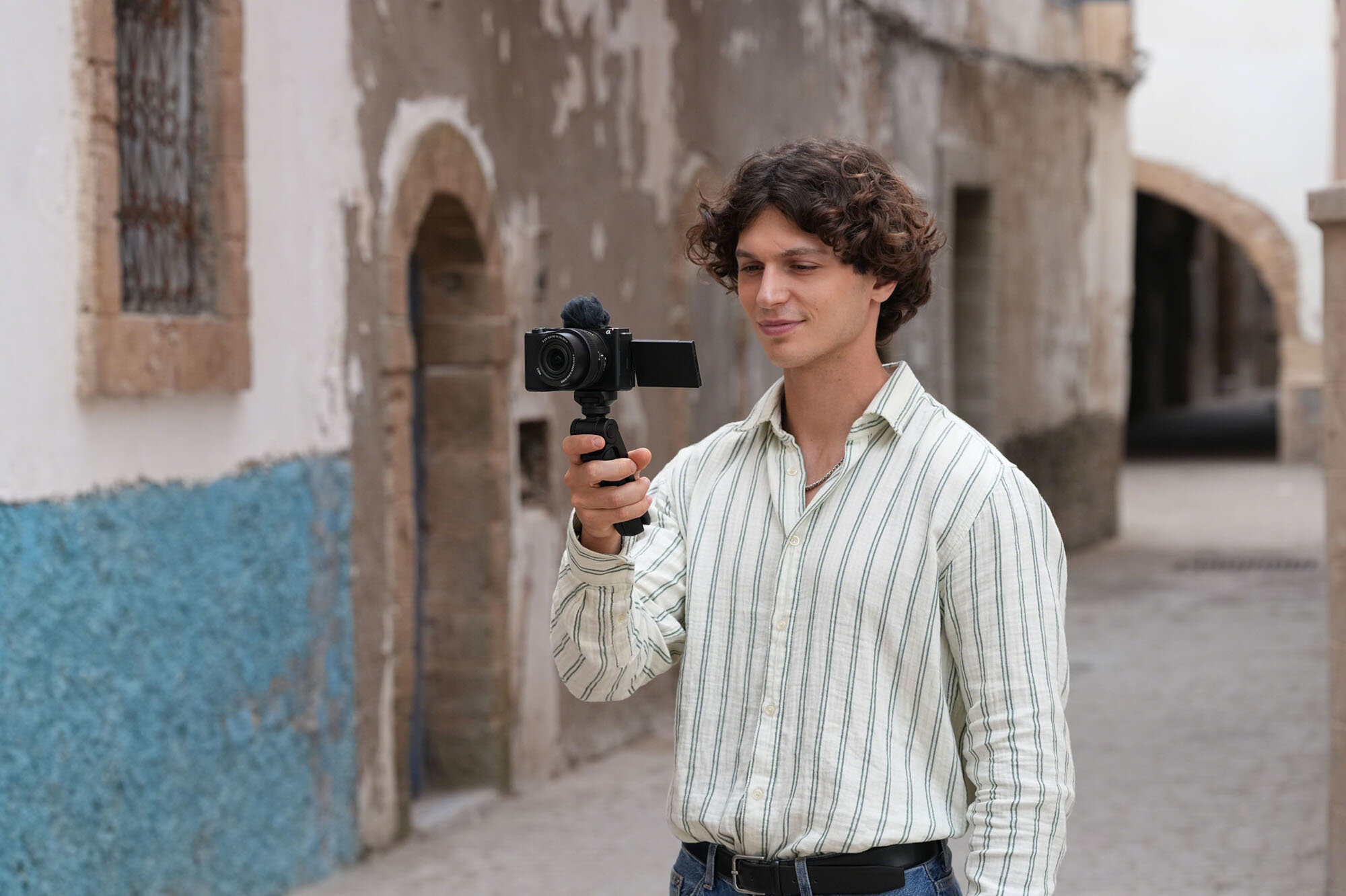 Mężczyzna w koszuli w paski filmuje kamerą na ulicy z kamiennymi budynkami.