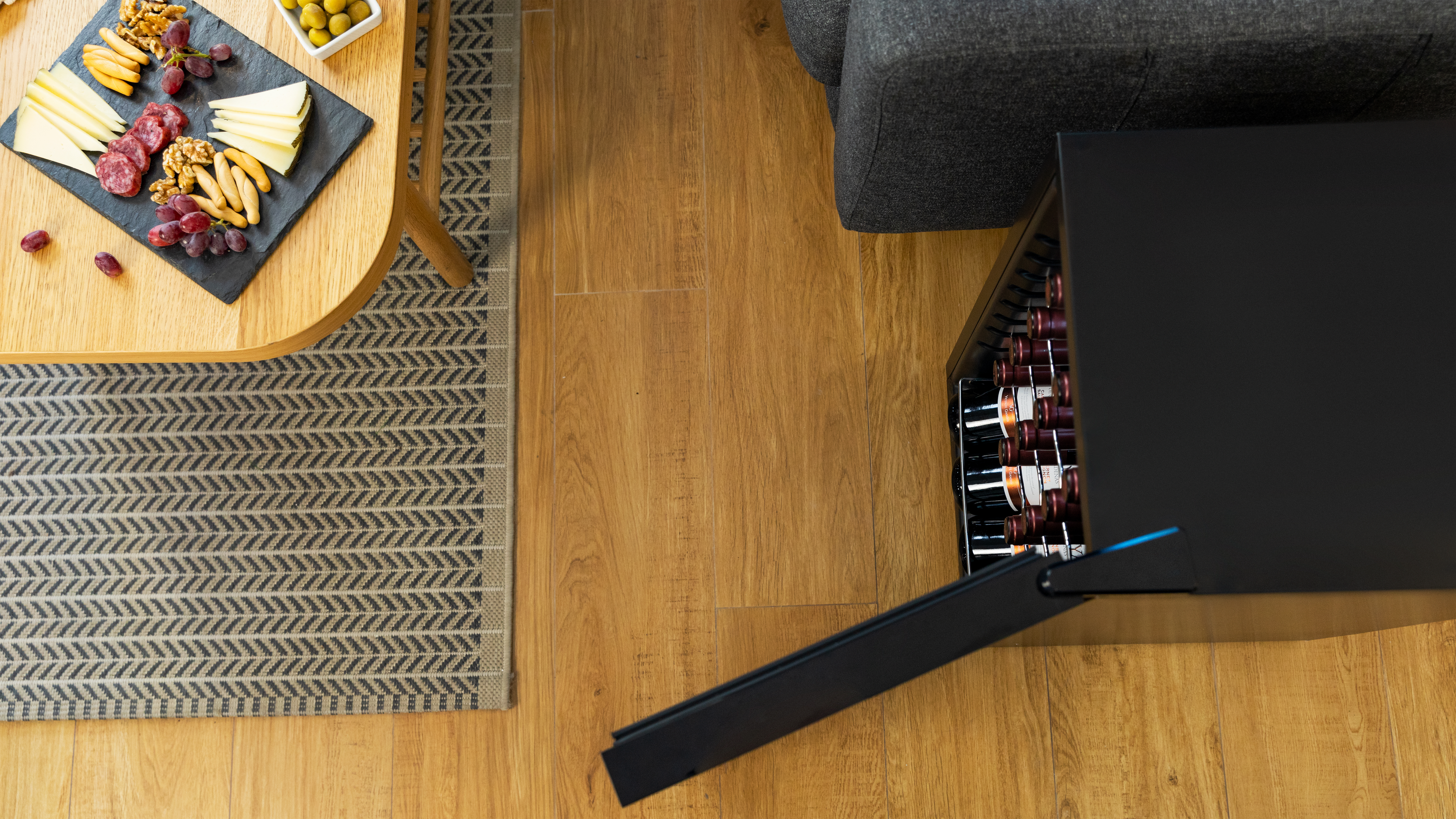 Mesa con aperitivos y nevera para vinos con puerta abierta mostrando botellas, sobre suelo de madera.