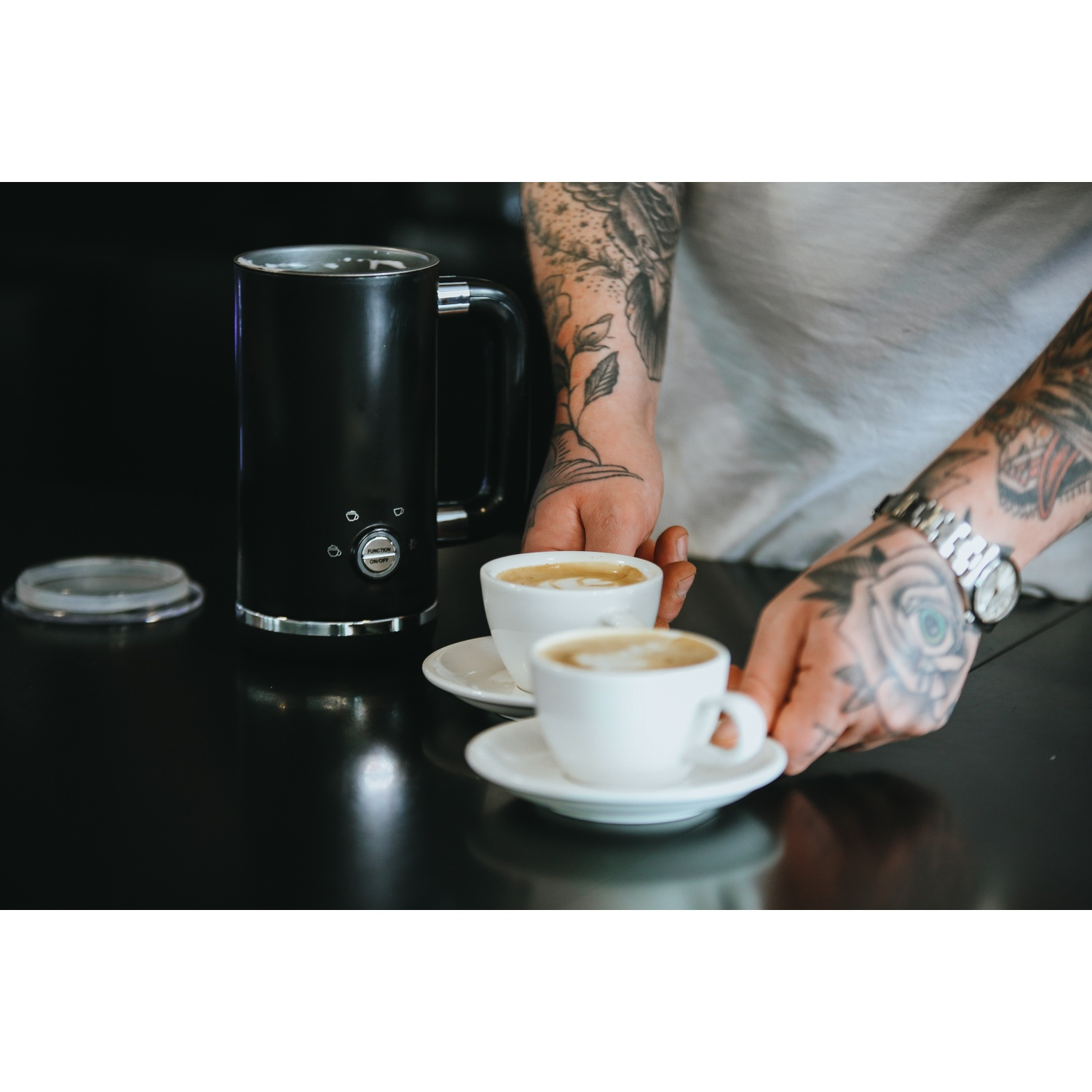 Una persona con tatuaggi sulle braccia serve due tazze di caffè. A sinistra c'è una macchina per il caffè nera.