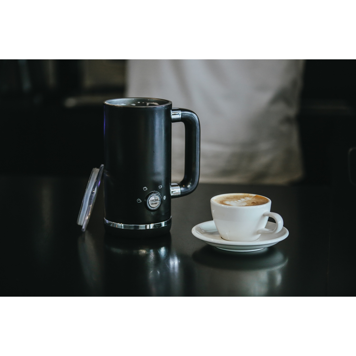 Montalatte nero e tazza di cappuccino su superficie scura. Persona sfocata sullo sfondo.