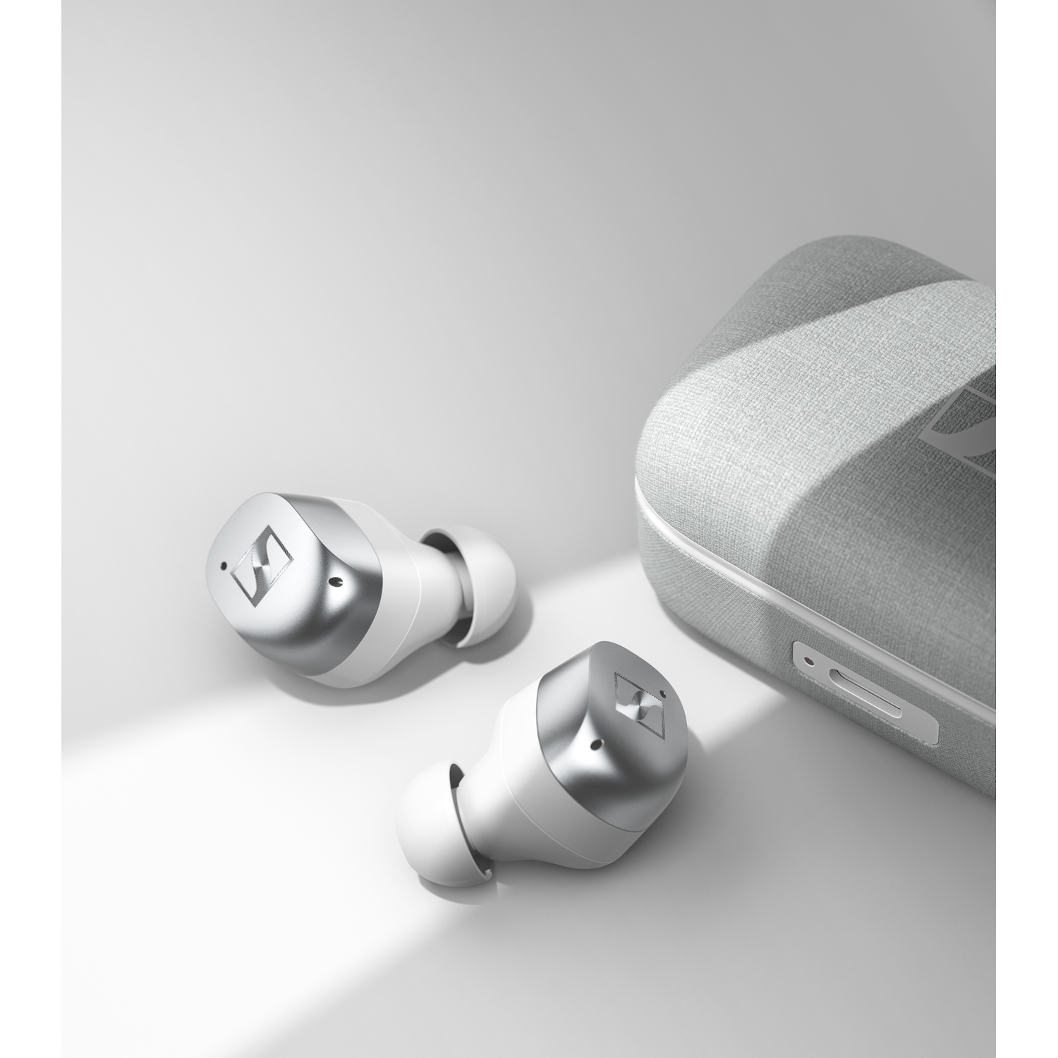 White Sennheiser earbuds and charging case on a white surface. The earbuds have silver accents.