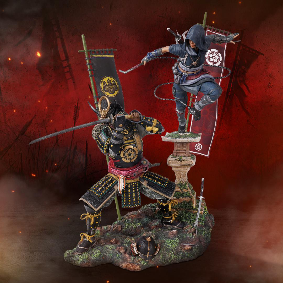 Statuen von zwei Samurai in schwarzer Rüstung, einer in Aktion. Rote Banner im Hintergrund, auf einer moosigen Basis.