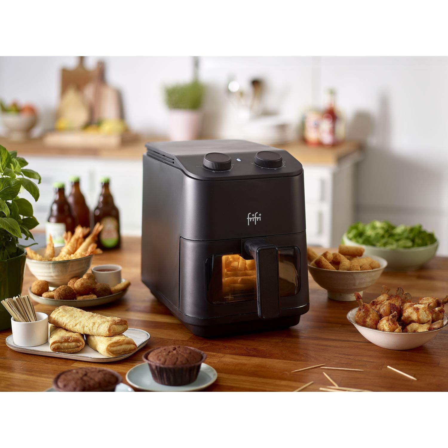 Zwarte airfryer met eten erin en eromheen op een houten tafel.
