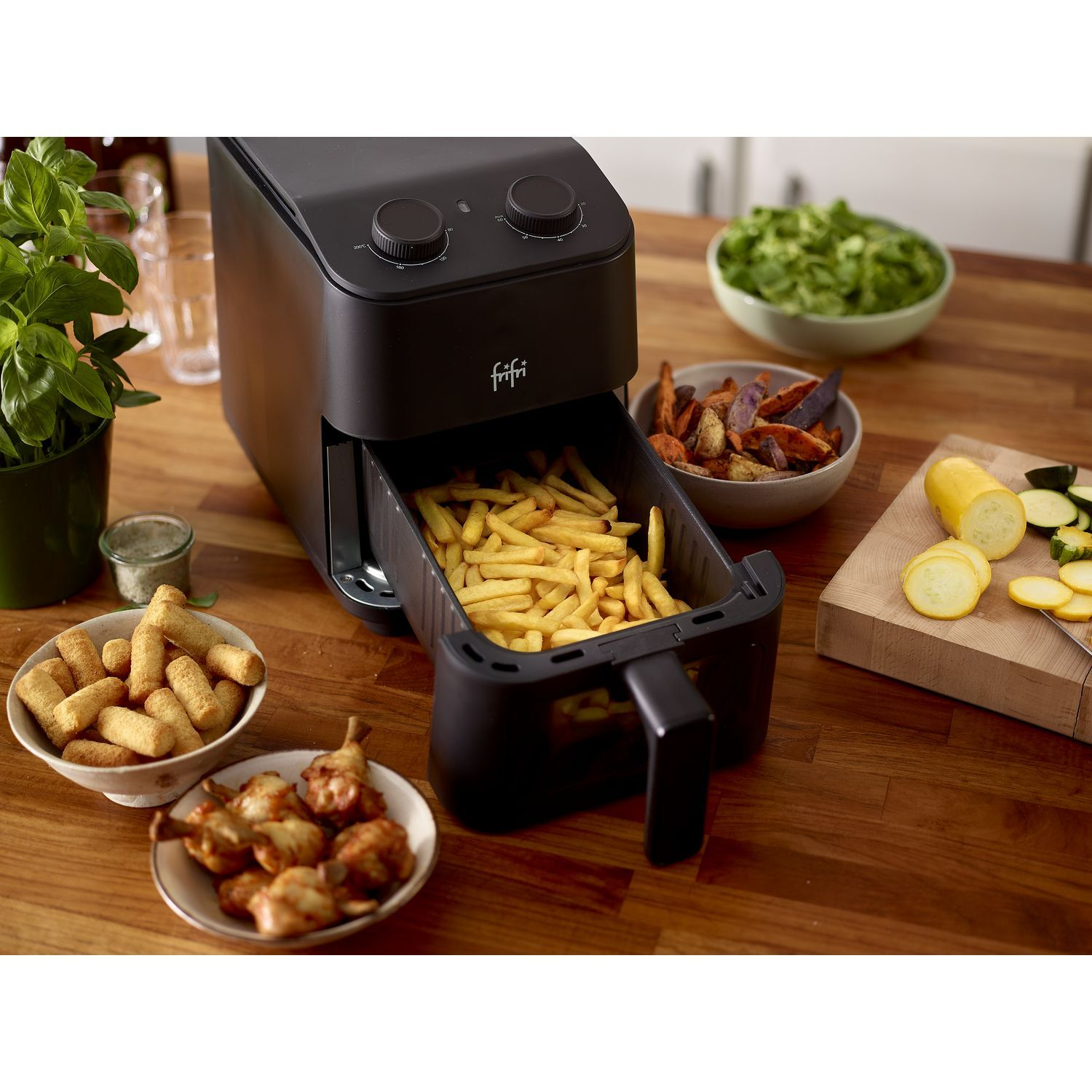 Zwarte airfryer met friet. Schalen met eten omringen de fryer. Een plant en een houten oppervlak.