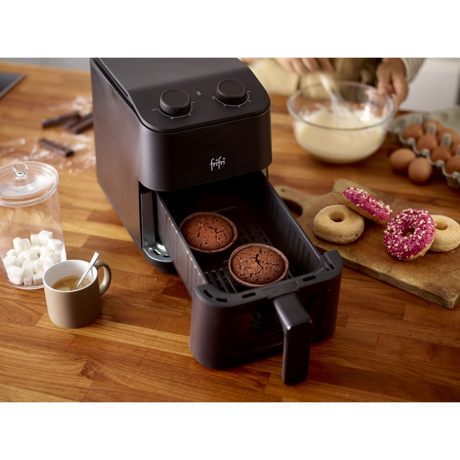 Een airfryer met twee cupcakes erin en donuts ernaast, met ingrediënten op de houten tafel.